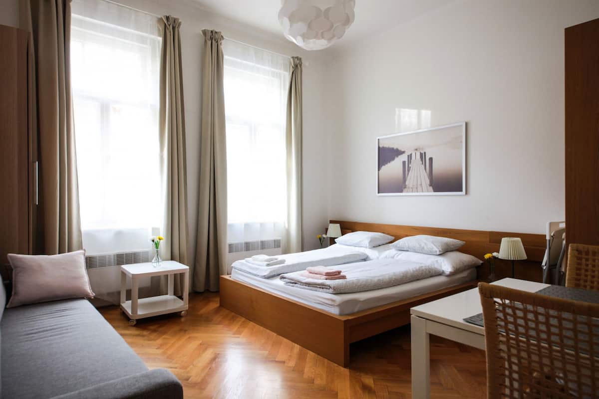 A hotel room with a bed, a sofa, a small table, a chair, a window with curtains, a wall-mounted picture, and a ceiling light fixture.