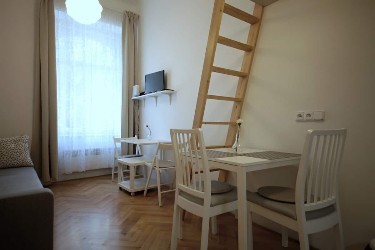A room with a wooden floor, a white desk with a computer, two white chairs, a small white shelf with a TV, a white sofa, and a window with sheer curtains.