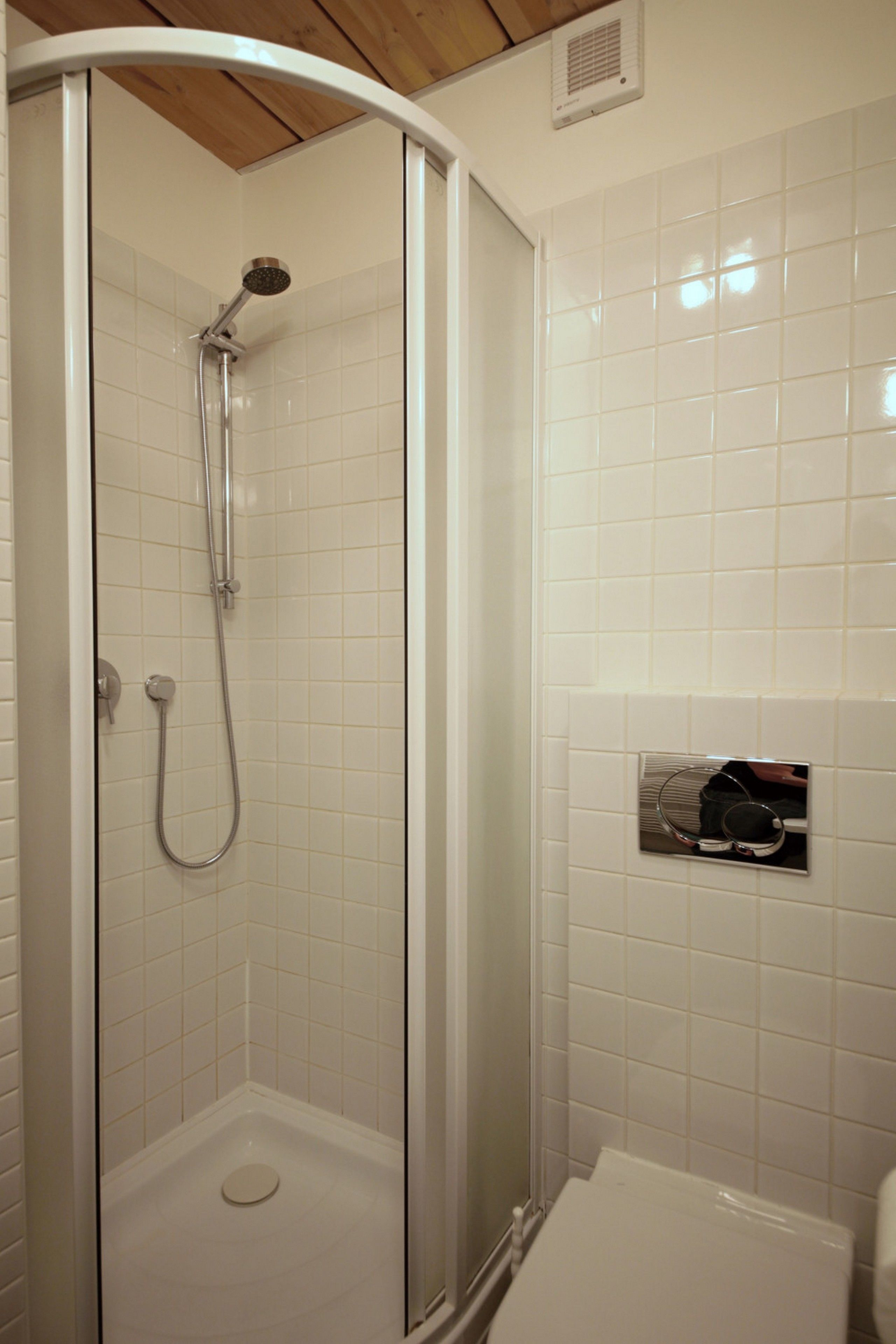 Une douche fermée par des parois en verre, dotée d’un pommeau de douche à main, d’un porte-serviettes mural et de toilettes, le tout dans une salle de bain aux murs recouverts de carreaux blancs.