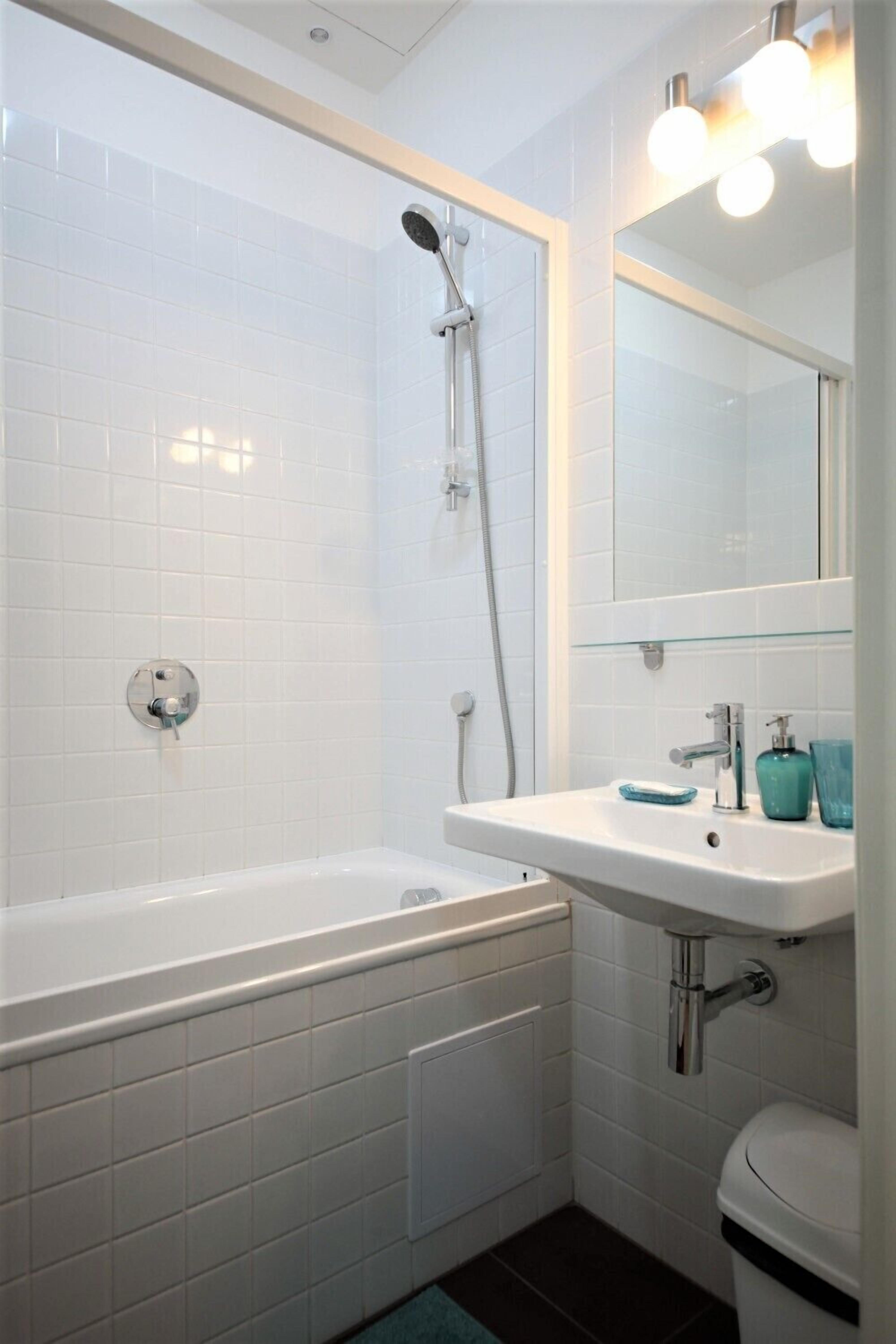 Un baño moderno con paredes de azulejos blancos, una bañera blanca, un lavabo con espejo y un inodoro.