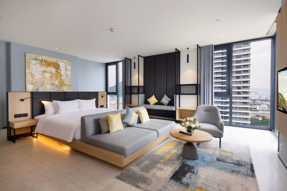 A modern hotel room with a large bed, a sofa, a coffee table, and a view of the city through large windows.