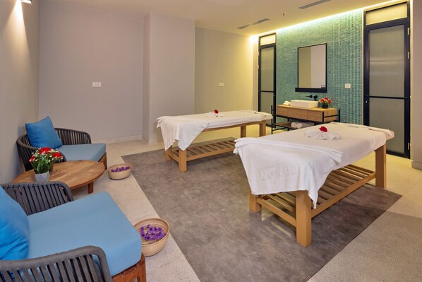 Un cuarto de spa con dos camillas de masaje, un sofá pequeño, una mesa de madera con un jarrón de flores y un lavamanos con espejo.