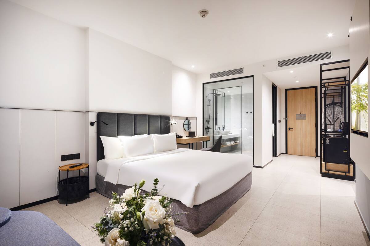 A modern hotel room with a large bed, bedside table, and a bathroom with a glass shower enclosure.