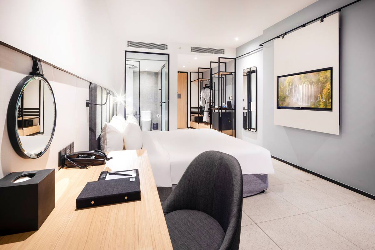 A modern hotel room with a large bed, a desk with a chair, a round mirror, and a framed picture on the wall.