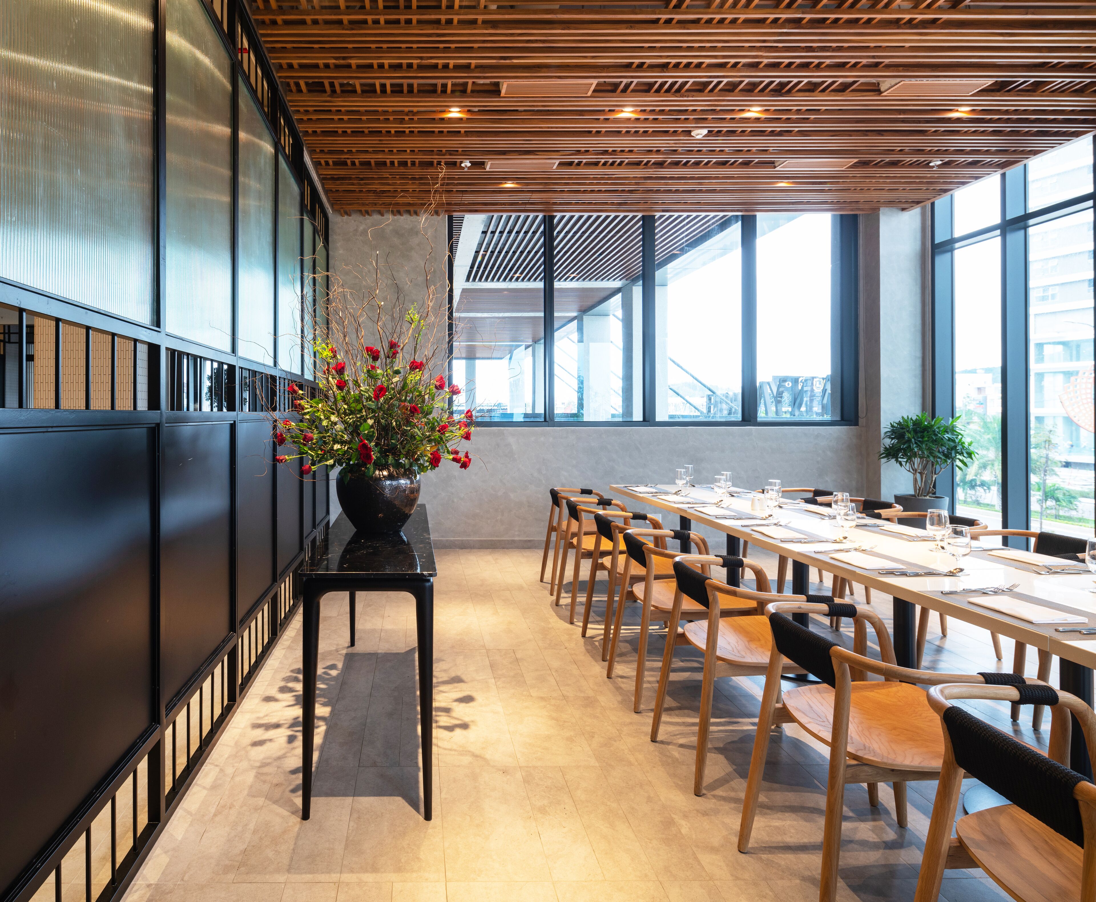 Una sala de conferencias moderna con una larga mesa, sillas de madera y una planta en maceta con flores rojas.