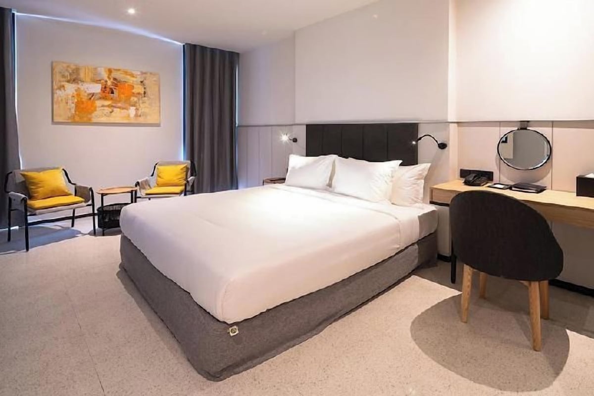 A modern hotel room with a large bed, a desk, a chair, and a bedside table.