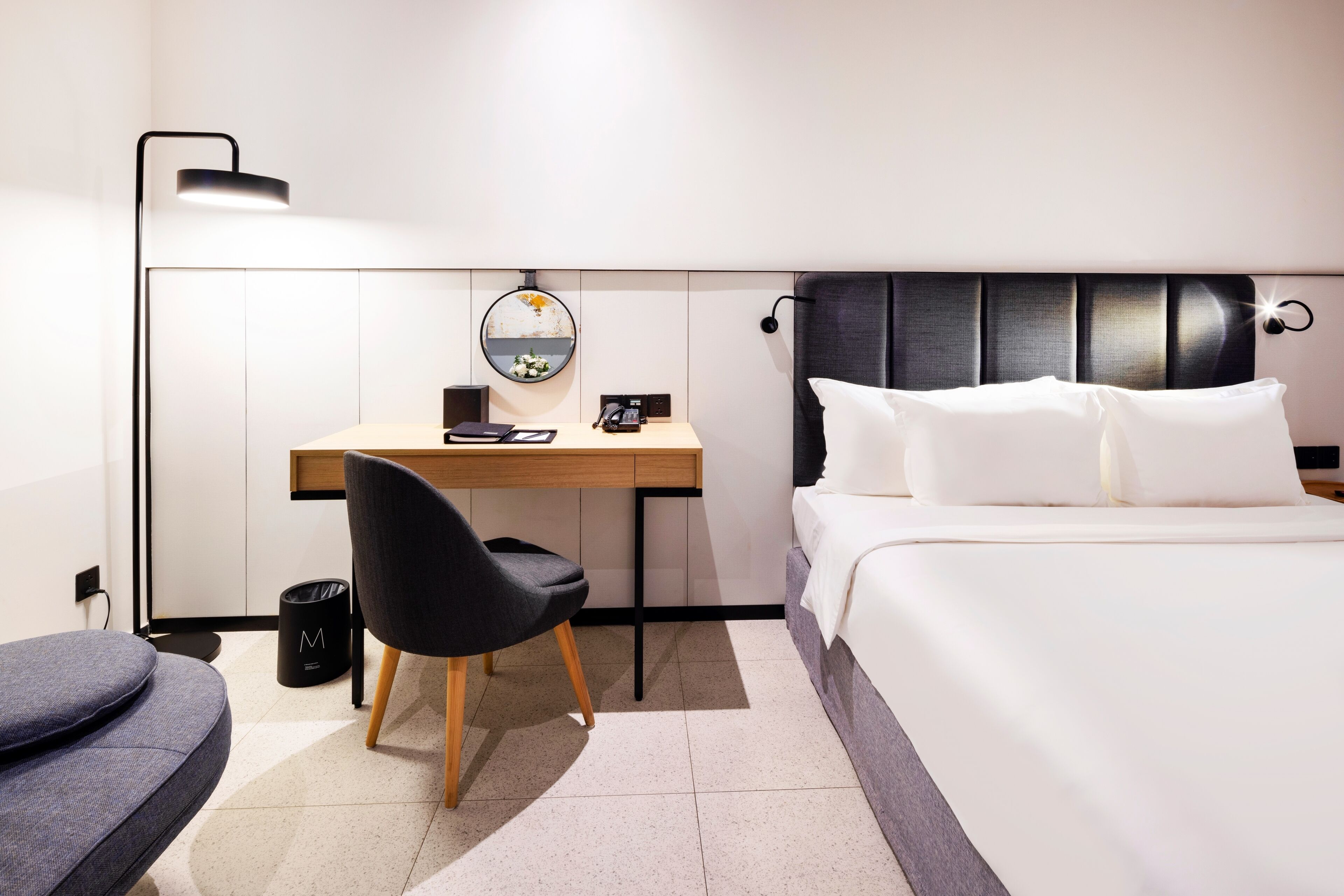 A modern hotel room with a bed, a desk, a chair, a lamp, and a trash bin.