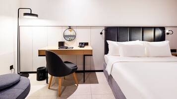 A modern hotel room with a bed, a desk, a chair, a lamp, and a trash bin.