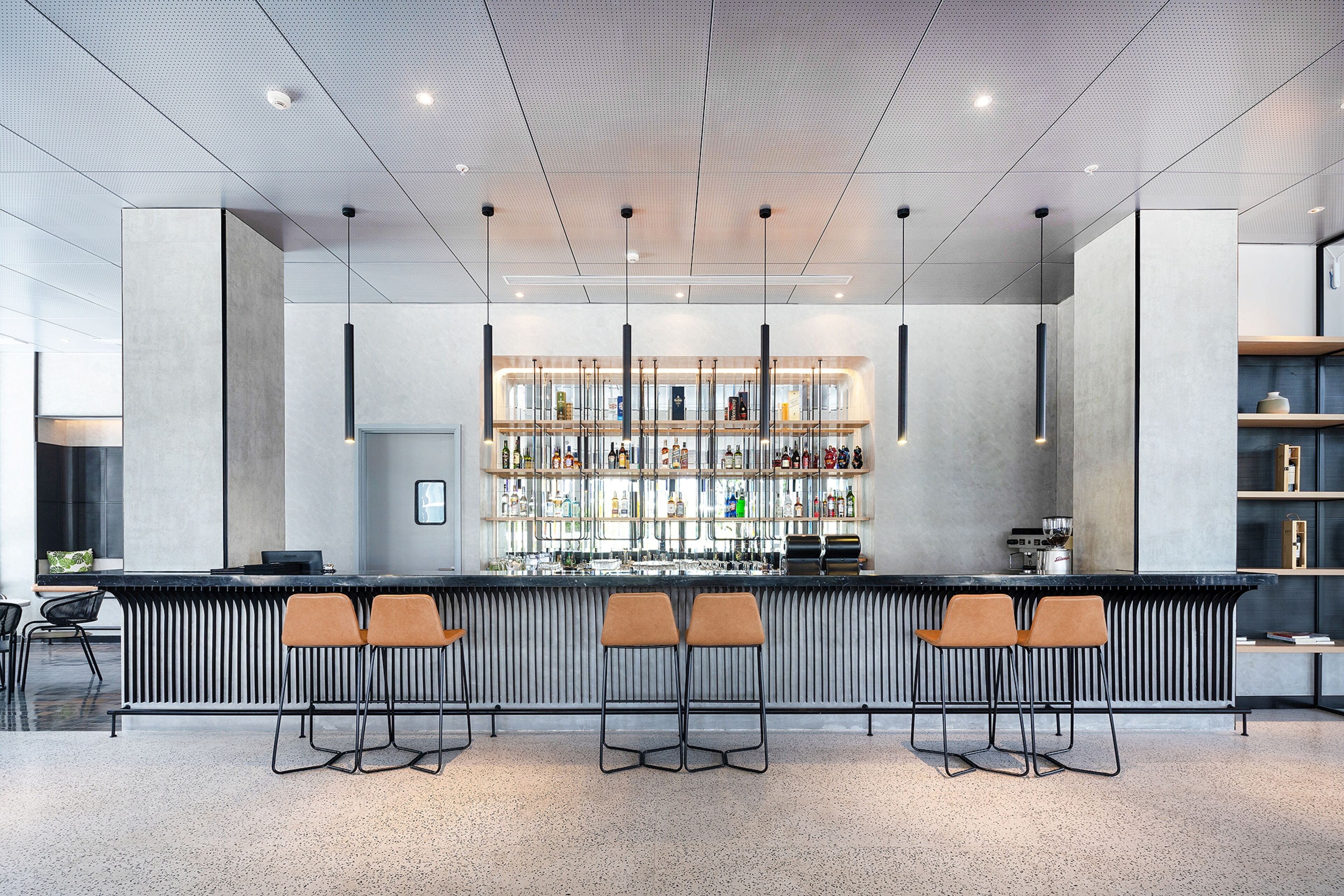 A modern bar area with a long counter, three bar stools, and a well-stocked shelf with various bottles.