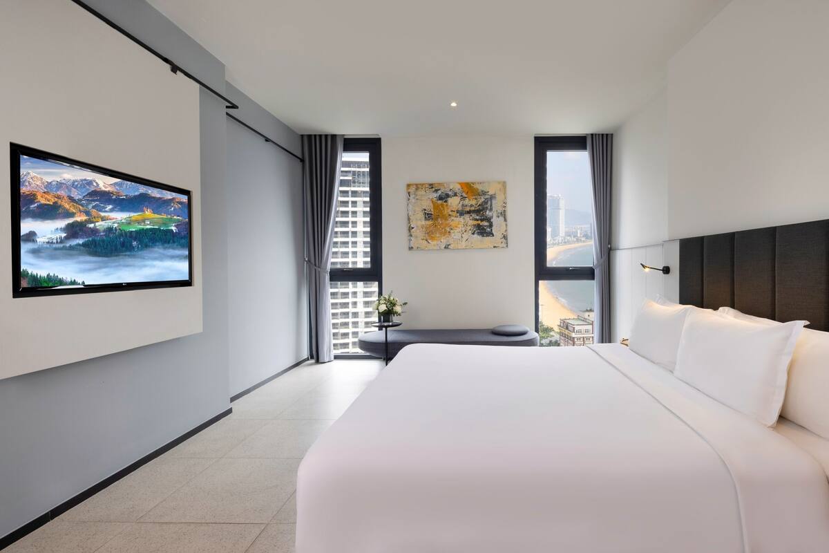 A modern hotel room with a large bed, a flat-screen TV mounted on the wall, a window with a view of a cityscape, and a painting on the wall.