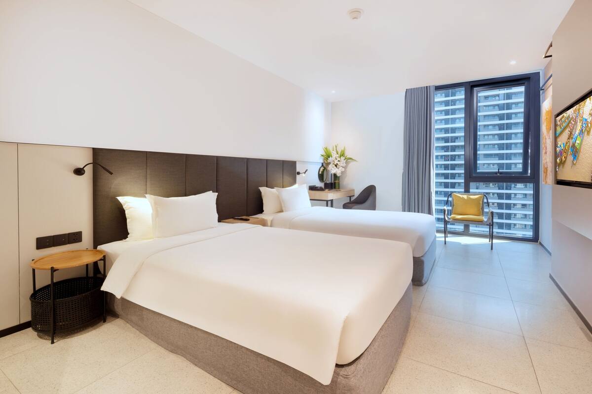 A modern hotel room with two beds, a desk, a chair, and a large window with a city view.