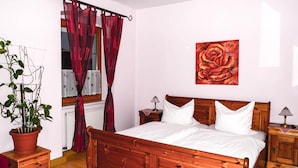 Ein hölzernes Bett mit weißer Bettwäsche, ein roter Vorhang, ein Blumentopf und ein Rosenbild an der Wand.