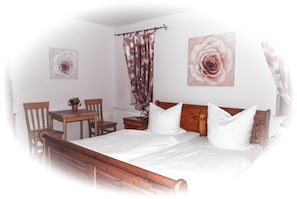 A bedroom with a wooden bed, white bedding, a wooden nightstand, a table with chairs, and two wall-mounted flower paintings.