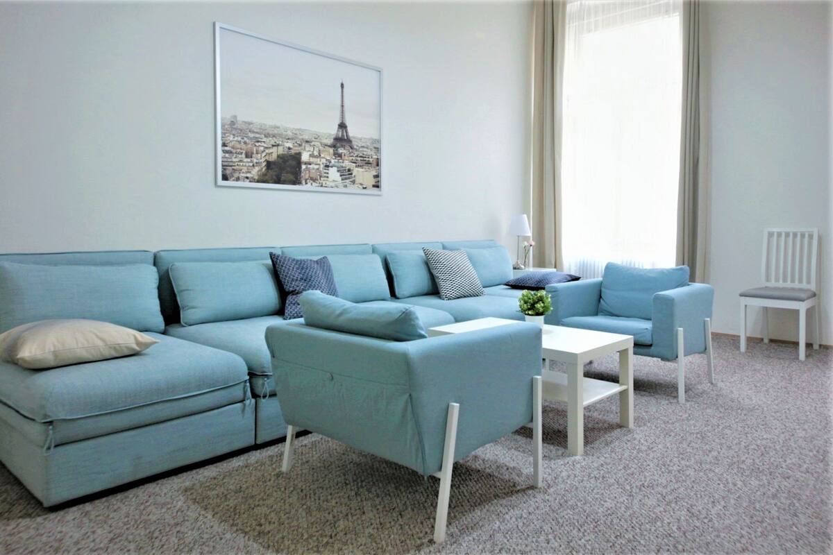 A living room with a light blue sofa, a white coffee table, and a white chair. There is a framed picture of a cityscape on the wall.