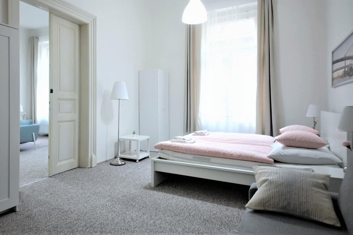 A bedroom with a bed, bedside lamps, a nightstand, a window with curtains, and a door leading to another room.