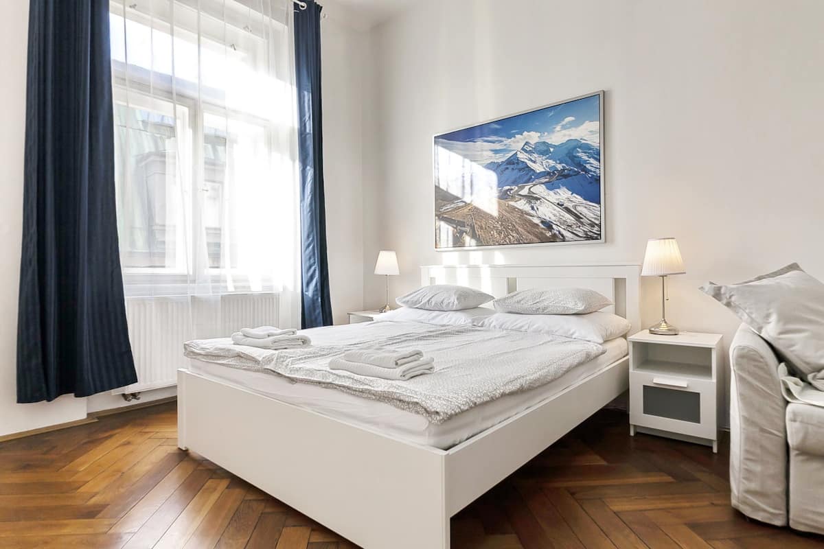 A modern hotel room with a large bed, two bedside tables, a nightstand, a window with curtains, and a mountain landscape painting on the wall.