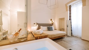 A modern hotel room with a large bathtub, a bed with patterned pillows, a bedside table with a bottle and glasses, and a mirror. - Suite1212 Pirrelli (Monopoli)