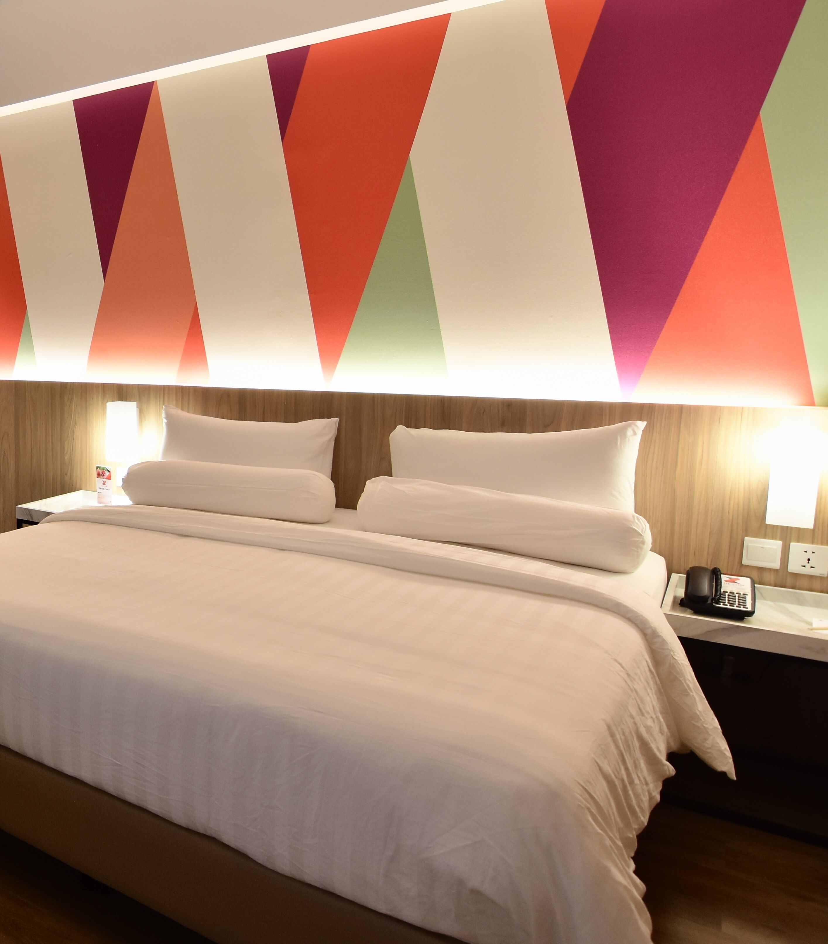 Una habitación de hotel con una cama grande, dos mesitas de noche y una pared decorada con un patrón geométrico de colores.