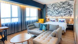 A modern hotel room with a sofa, a round table, a bed with colorful pillows, and a large abstract wall art piece. - The Zuri Dumai (Dumai)