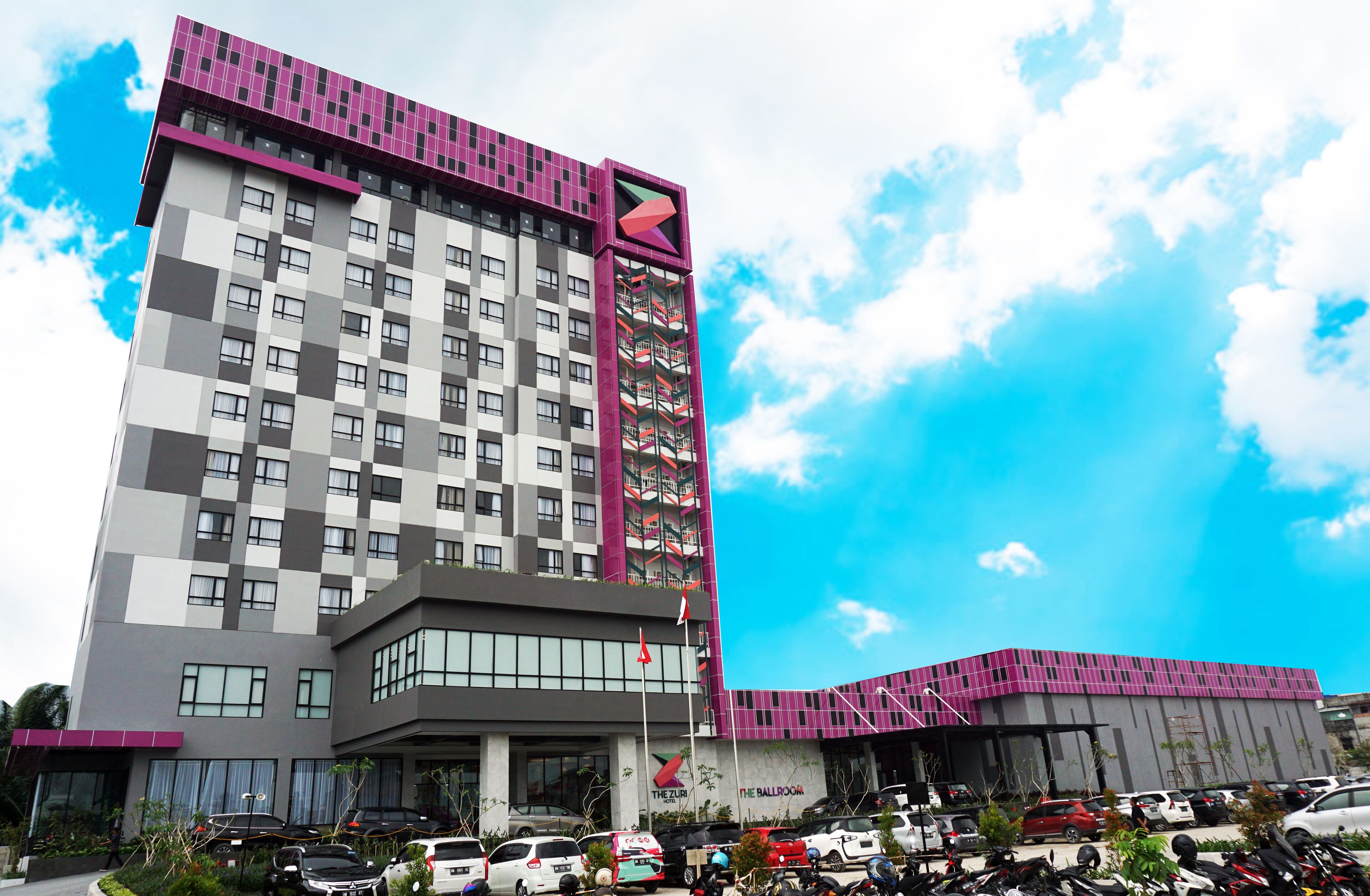 Un hôtel moderne à la façade à motifs géométriques, doté de nombreux balcons et disposant d’un parking où sont stationnés divers véhicules.