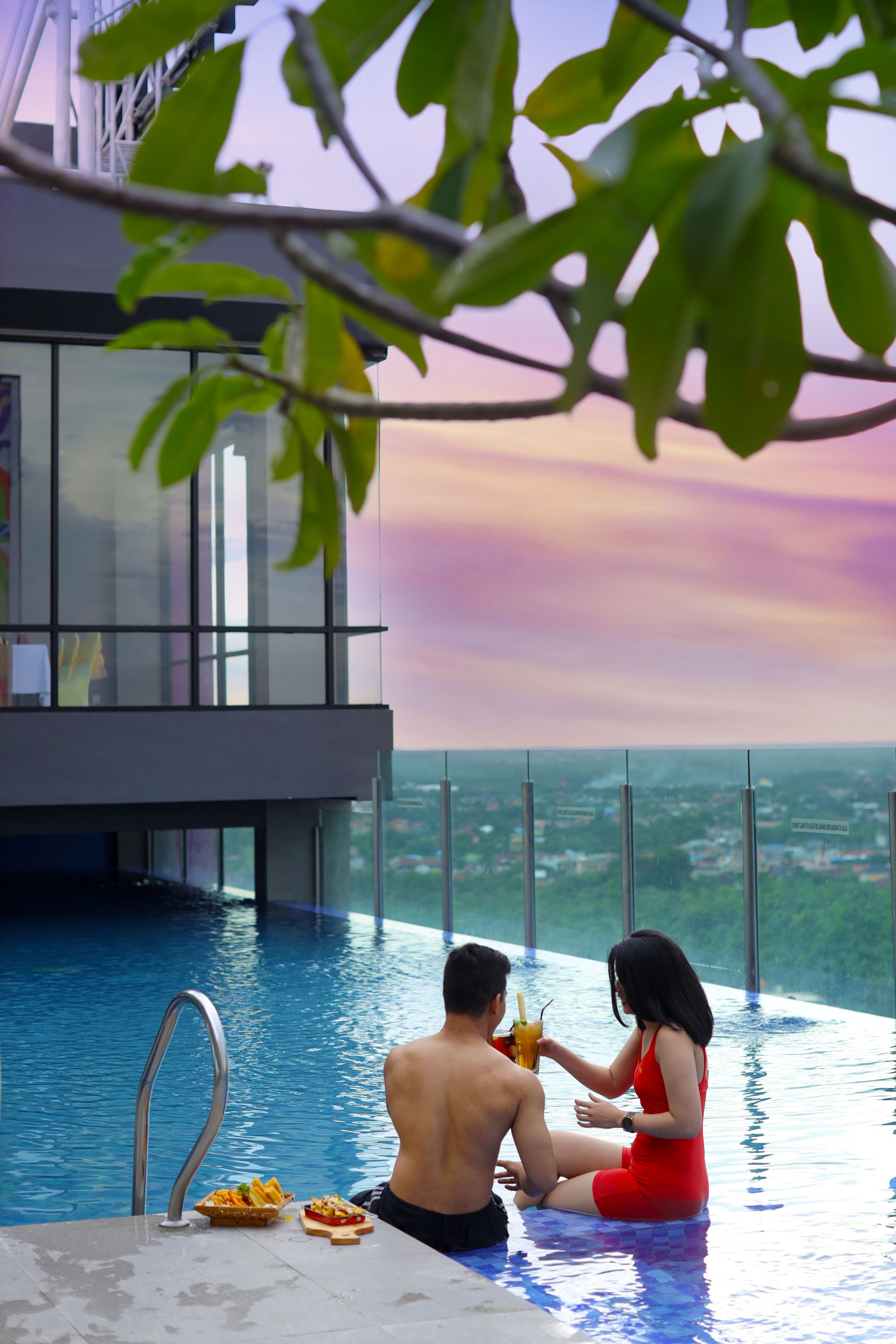 Una pareja sentada junto a una piscina infinita con vistas a la ciudad, un edificio moderno con ventanas de vidrio y un cielo despejado.