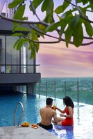 A couple sitting by an infinity pool with a city view, a modern building with glass windows, and a clear sky.