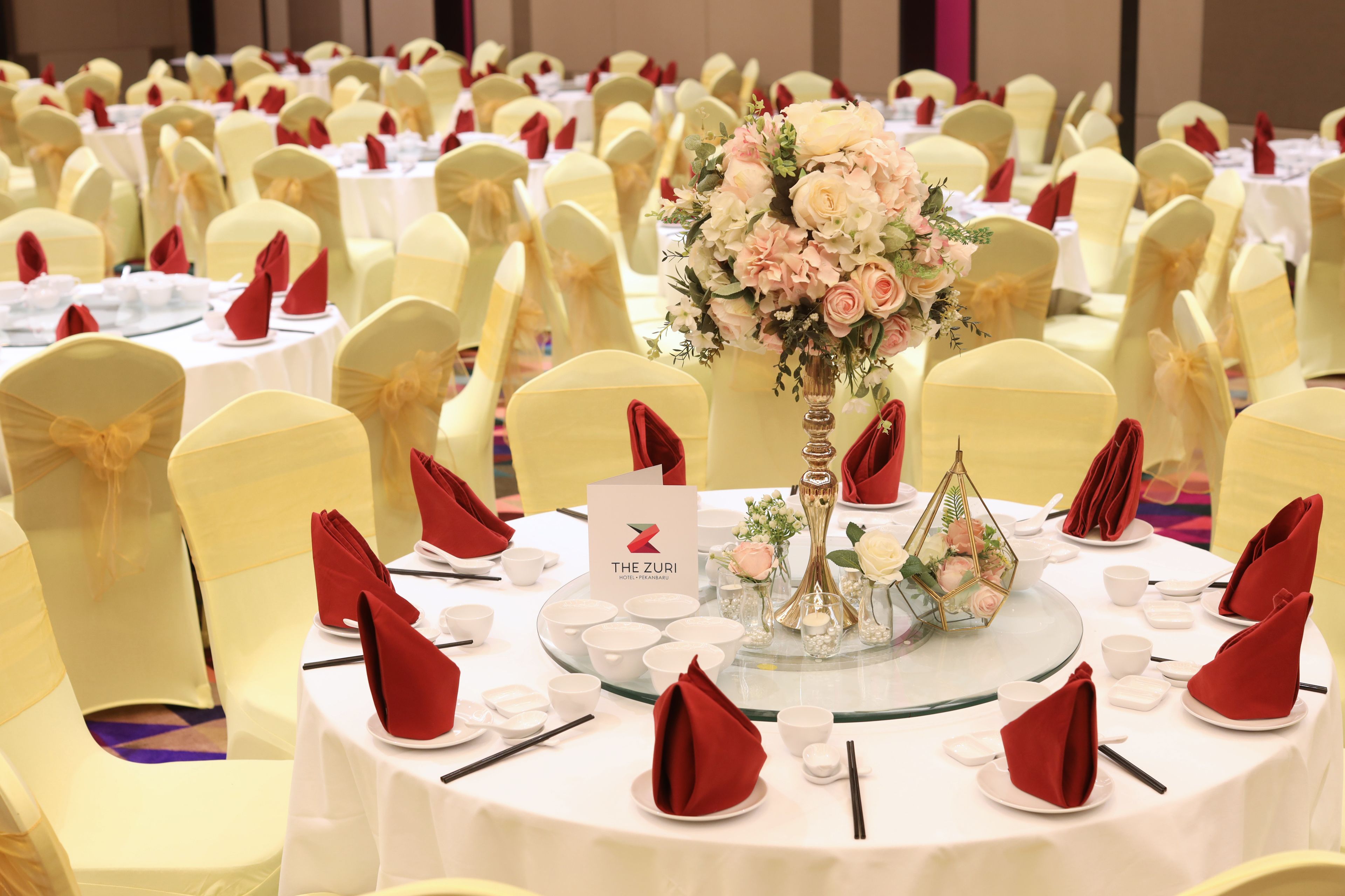 A banquet hall with round tables set for a formal event, featuring white tablecloths, red napkins, and floral centerpieces.