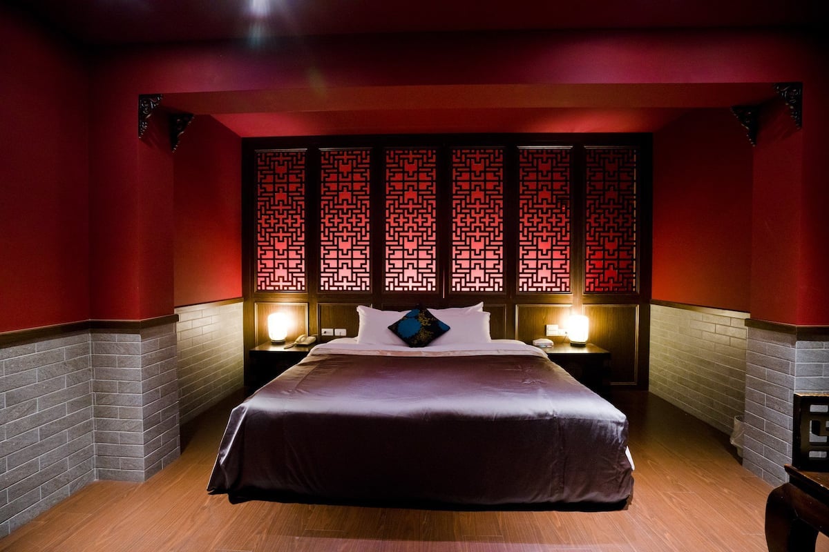 Una habitación de hotel con una cama grande, dos mesitas de noche con lámparas y un separador decorativo con un motivo geométrico.