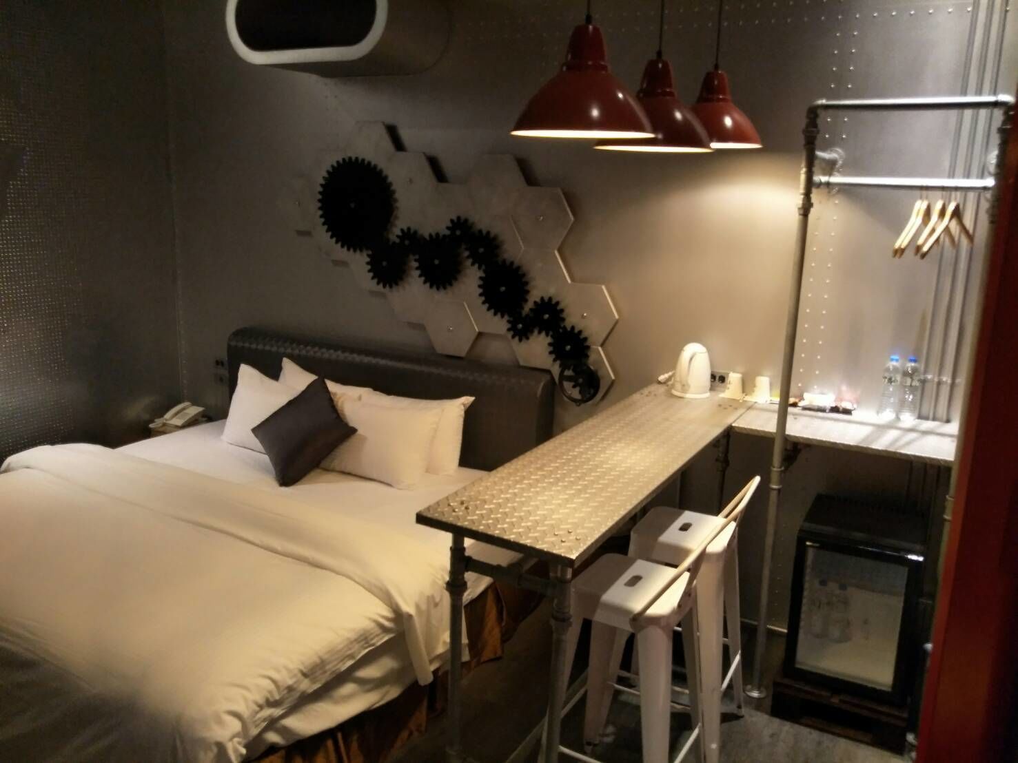 Quarto de hotel compacto com uma cama, uma secretária pequena e uma cadeira. Há três luminárias pendentes sobre a secretária.