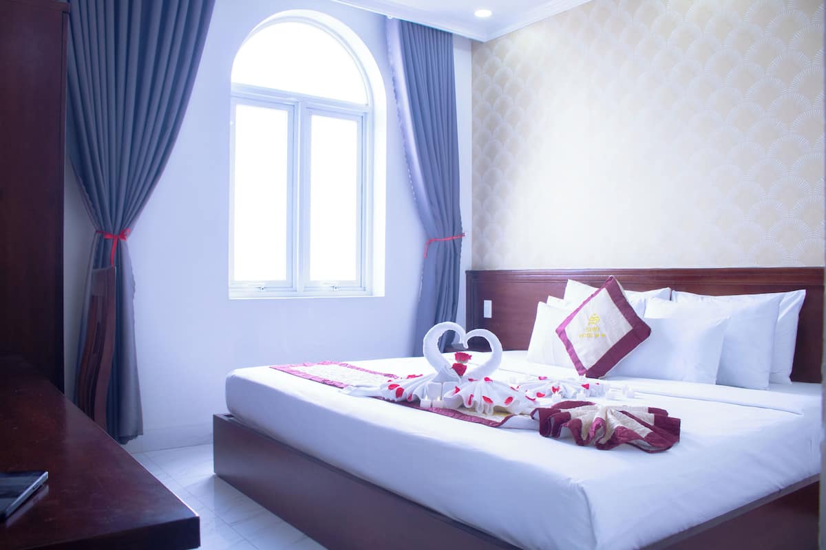 A hotel room with a bed, curtains, a window, and a headboard with a pillow.