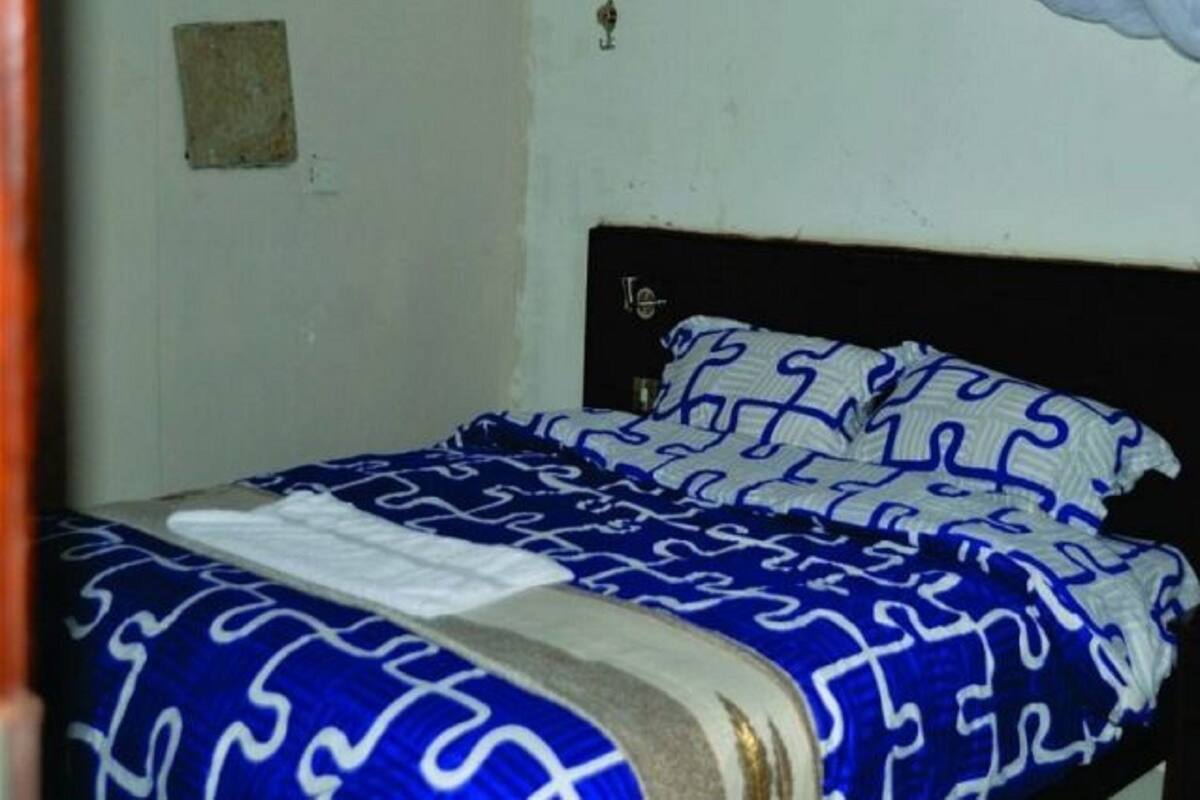 Uma cama de solteiro com colcha e almofadas estampadas, cabeceira de madeira e uma pequena janela quadrada na parede.