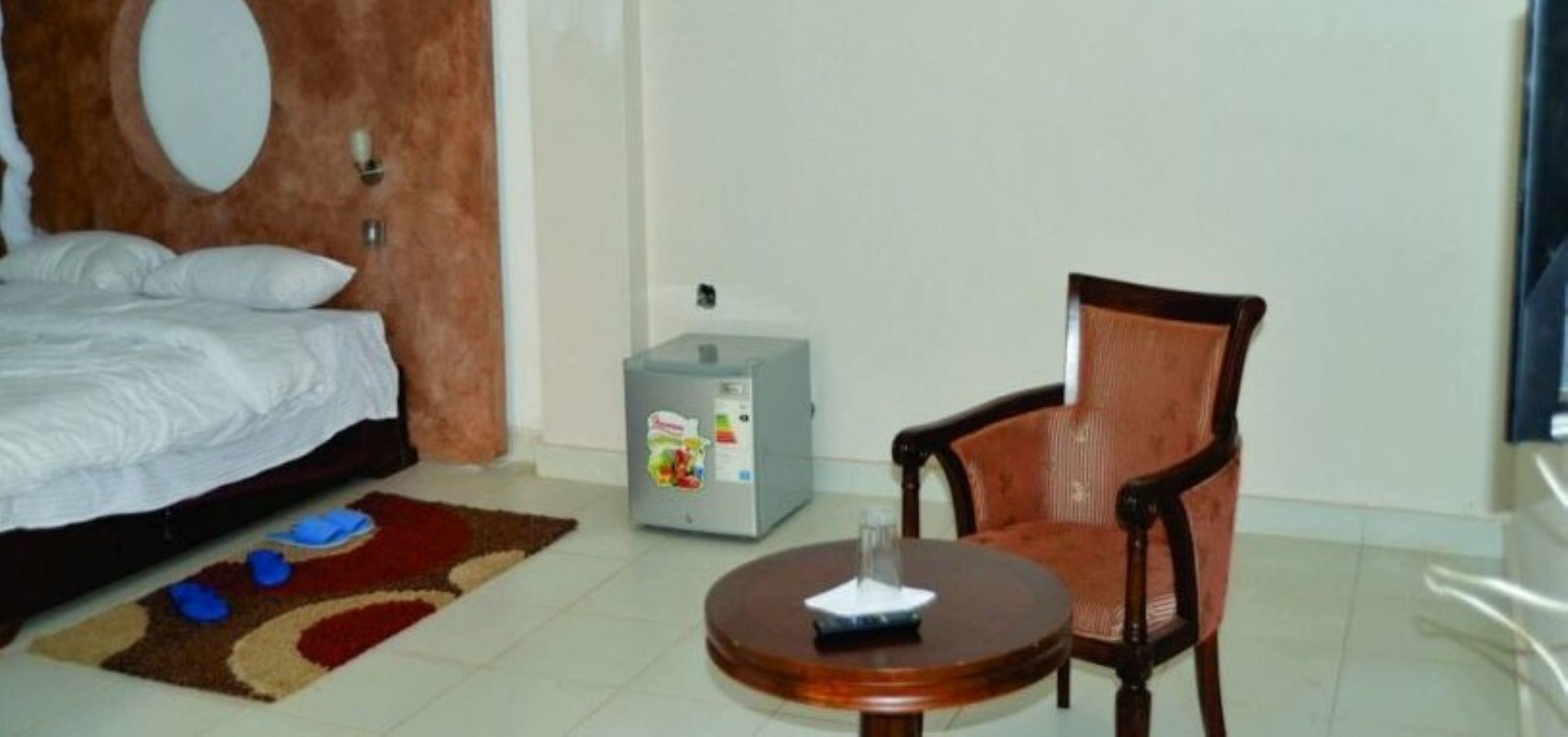 A bedroom with a bed, a chair, a small table, and a refrigerator.