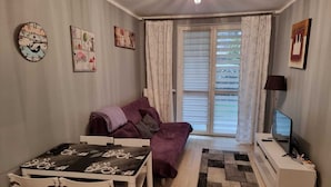 A cozy living room with a sofa, a dining table, and a TV. There are curtains and a window with blinds.