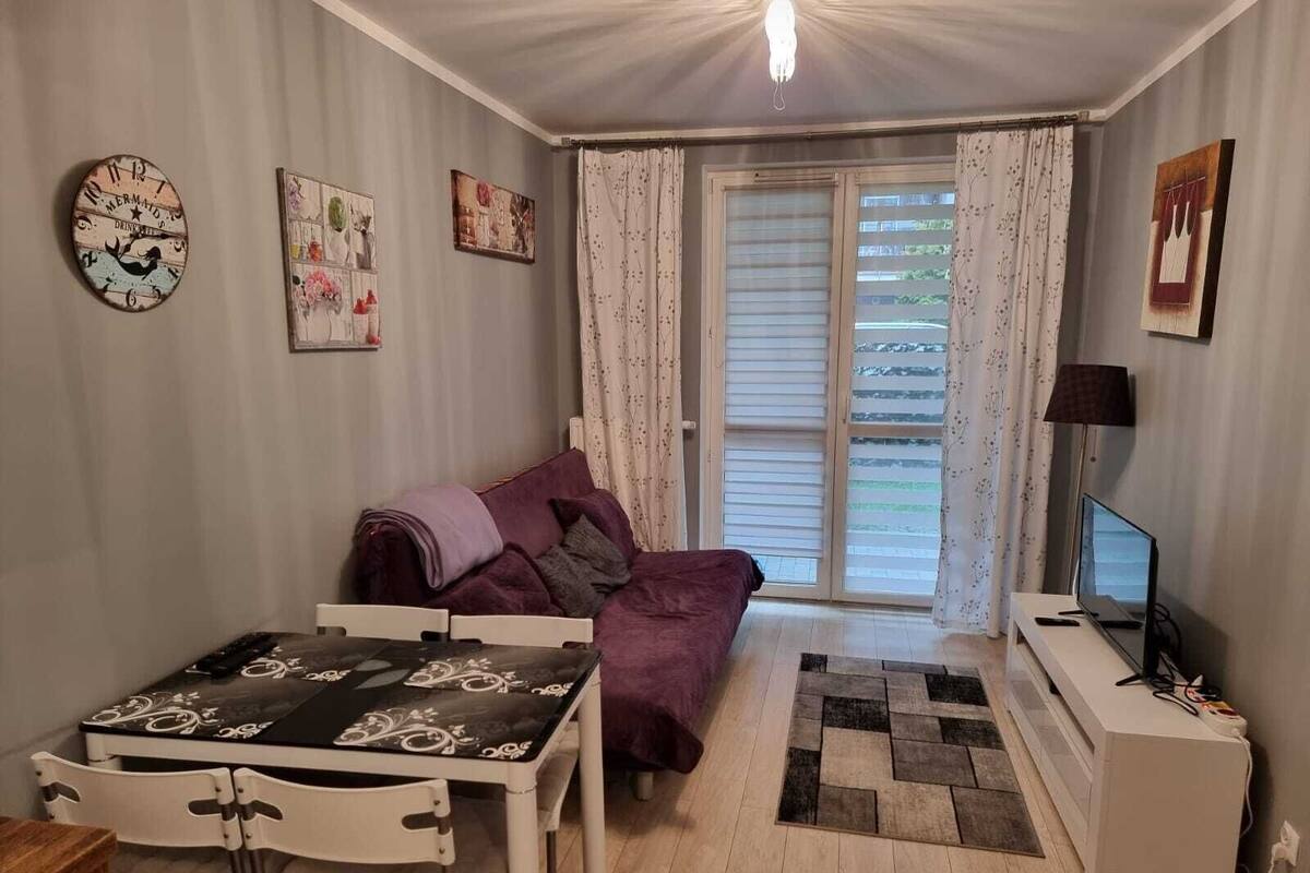 Un salon confortable avec un canapé, une table à manger et un téléviseur. Il y a des rideaux et une fenêtre avec des stores.