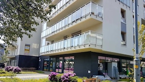 A modern multi-story building with balconies, a ground-level shop, and a well-maintained garden with various plants.
