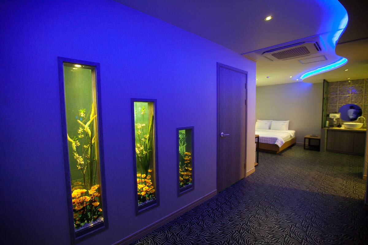 Habitació d'hotel amb llit, taula de nit petita i un altre taulell. Hi ha tres finestres verticals amb decoracions florals.