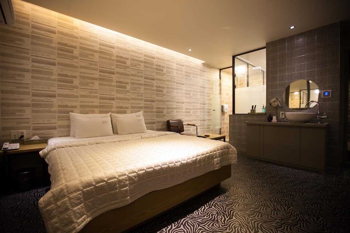 Habitació d'hotel moderna amb un llit gran, una tauleta de nit, un escriptori, un bany amb un lavabo i un mirall, i un paviment amb disseny.
