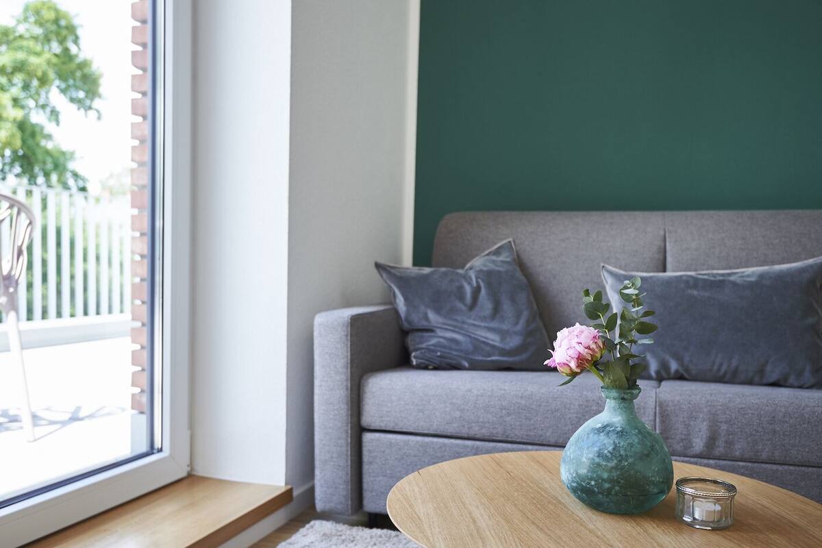 Una sala de estar moderna con un sofá gris, una mesa de centro de madera, un florero con flores y vistas al exterior a través de una ventana.