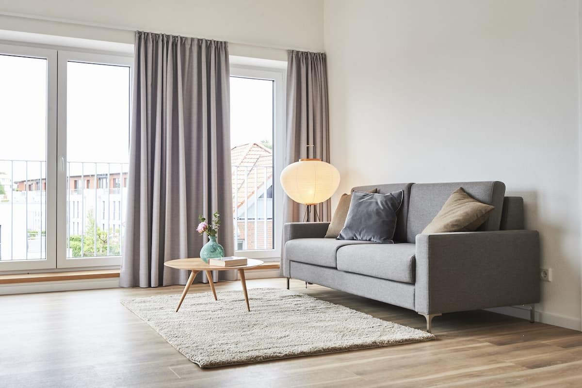 Una sala de estar moderna con un sofá gris, una mesa de centro redonda, un jarrón con flores y un ventanal con cortinas.