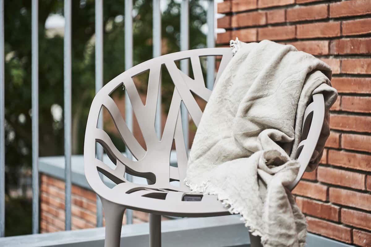 Una silla metálica blanca con cojín, ubicada en un balcón con un muro de ladrillo al fondo.
