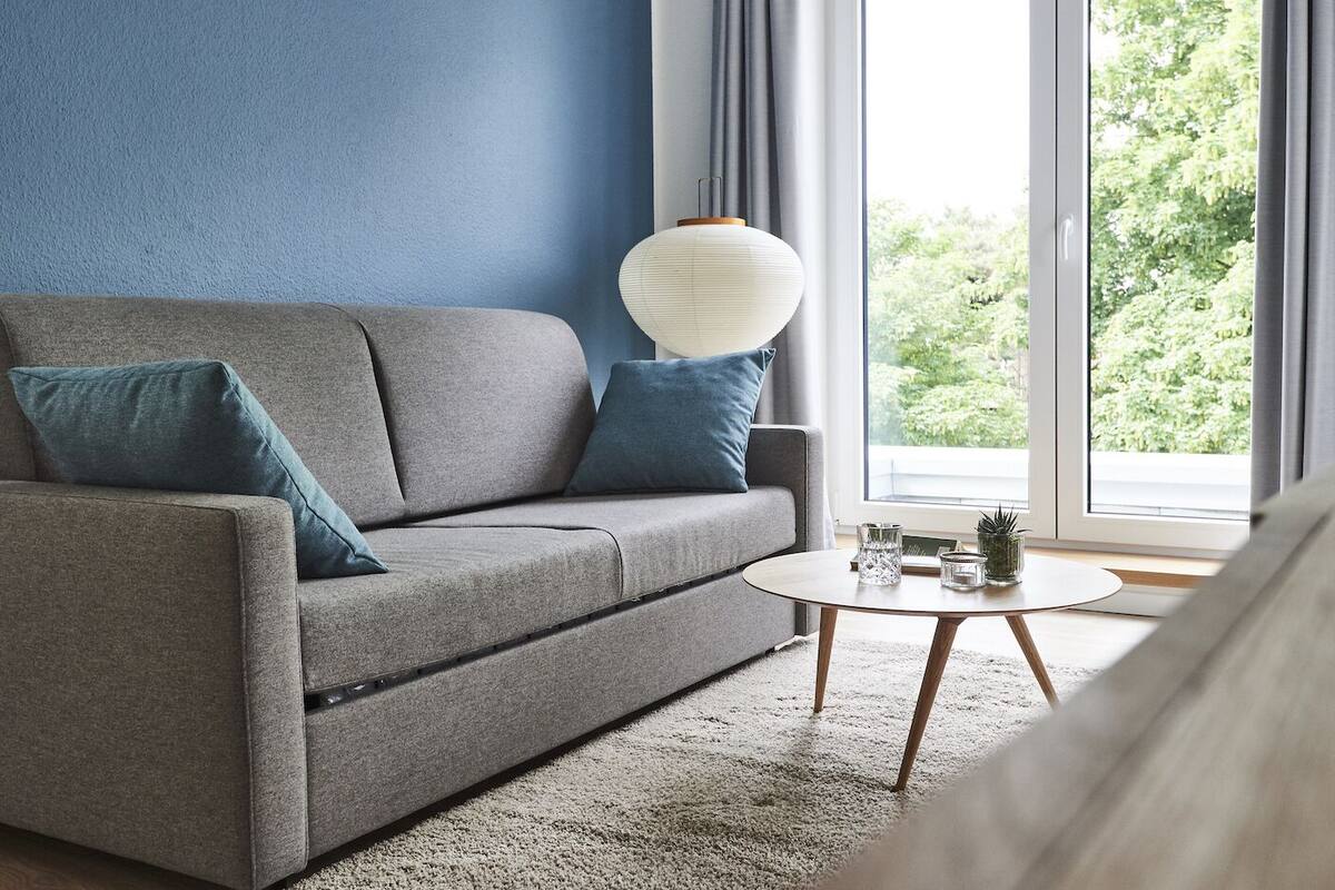 Una sala de estar moderna con un sofá gris, dos cojines azules, una mesa de centro redonda y una lámpara blanca.