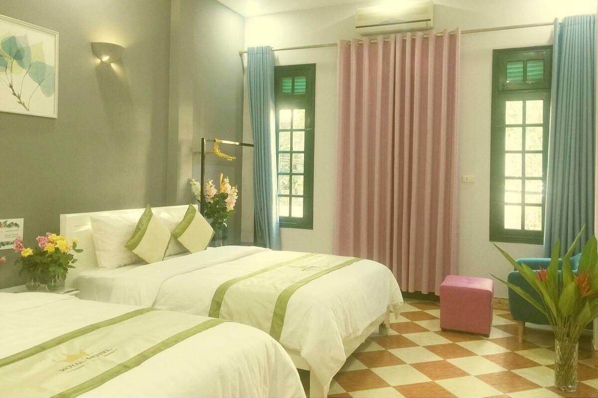 A hotel room with two beds, a small table with flowers, a chair, and a window with curtains.