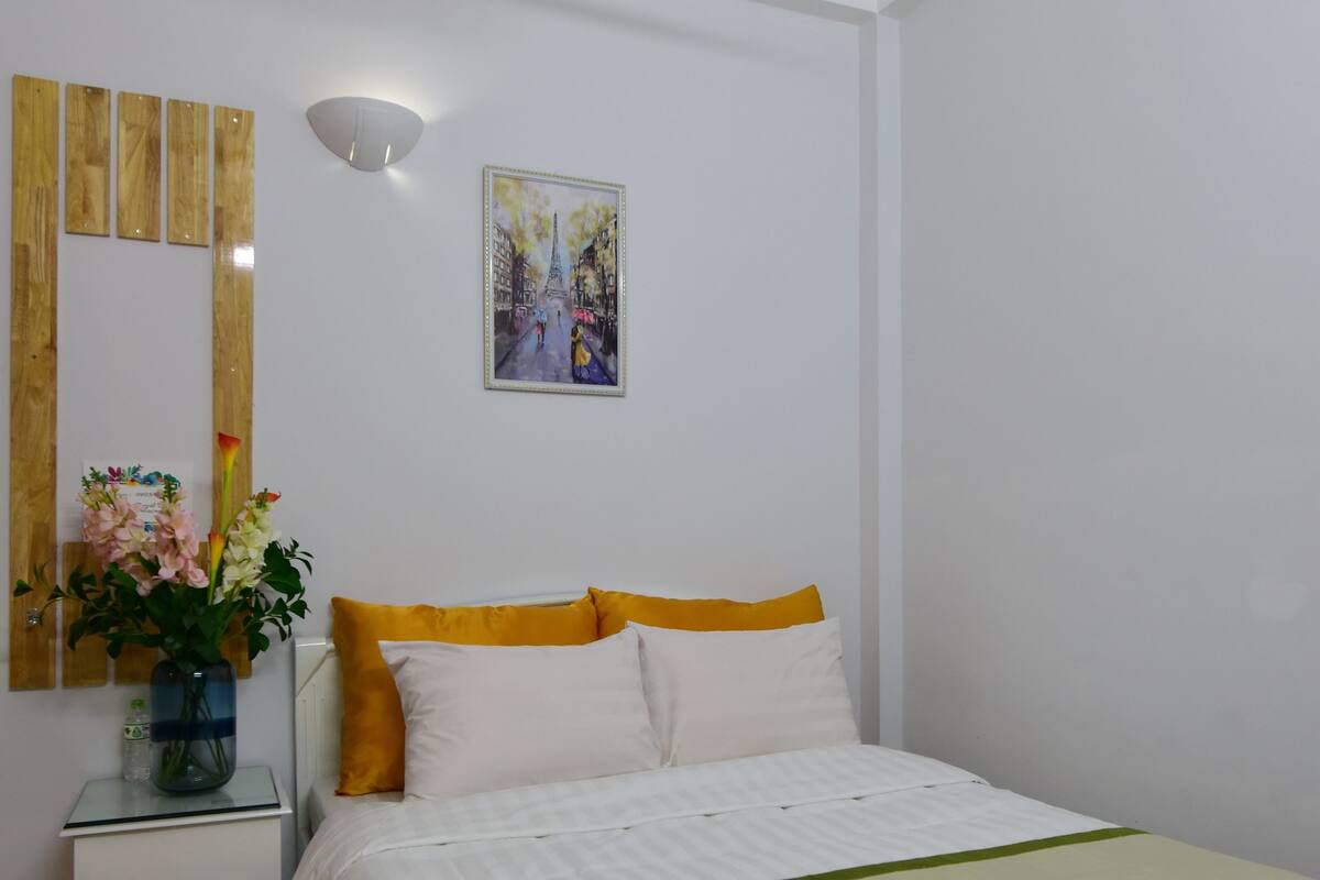 A hotel room with a bed, a nightstand with a vase of flowers, a painting on the wall, and a checkered floor.