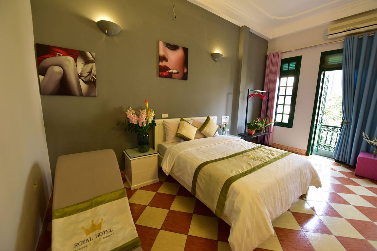 A hotel room with a checkered floor, a bed with a green and white bedspread, a nightstand with a vase of flowers, and a wall with two framed pictures.