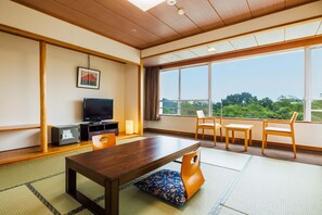 Une pièce avec un plafond en bois, une grande fenêtre donnant sur un paysage verdoyant, un téléviseur et un coin repas avec des tatamis.