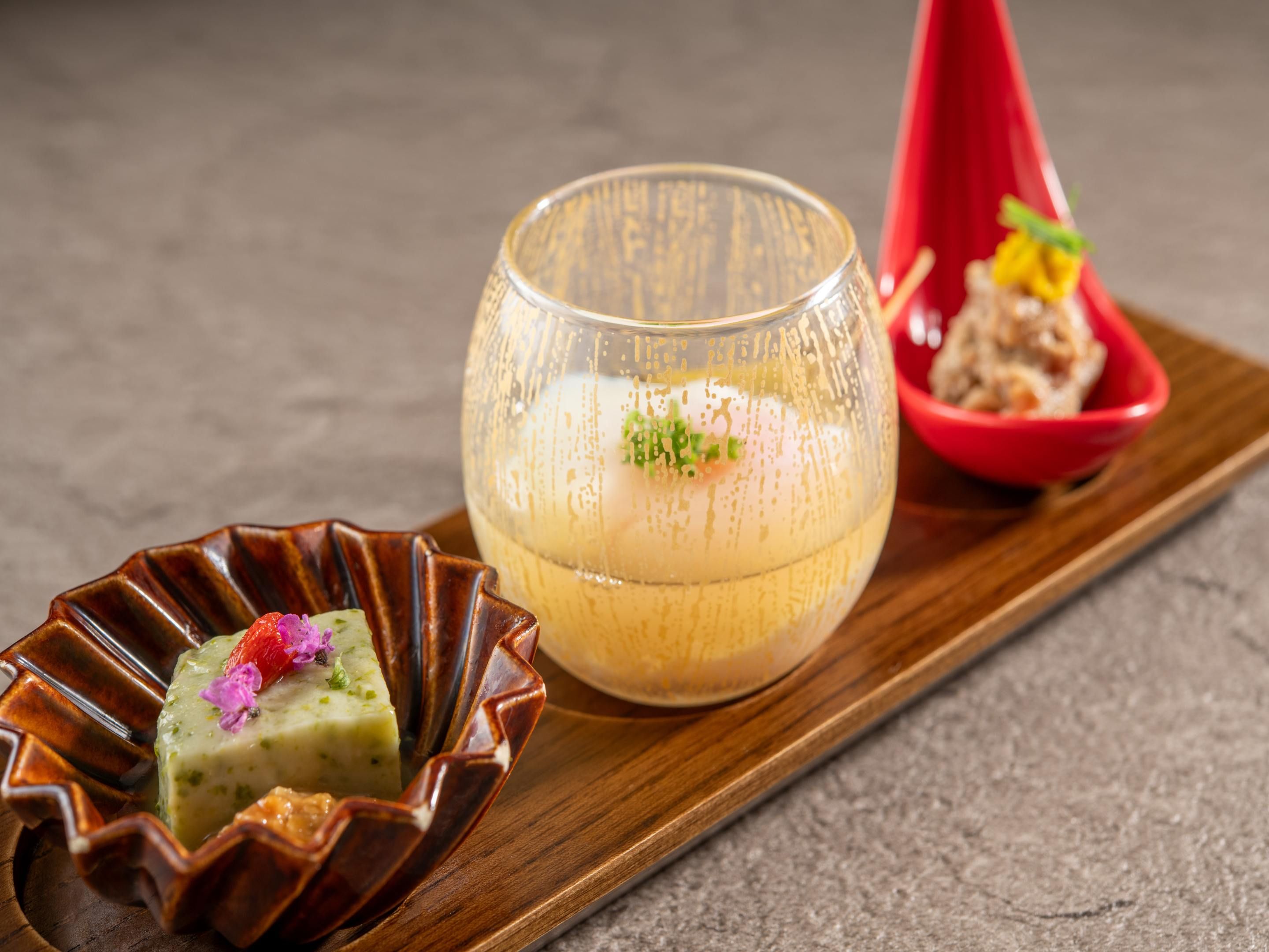 A dessert presentation with a glass, a red bowl, and a brown dish on a wooden board.