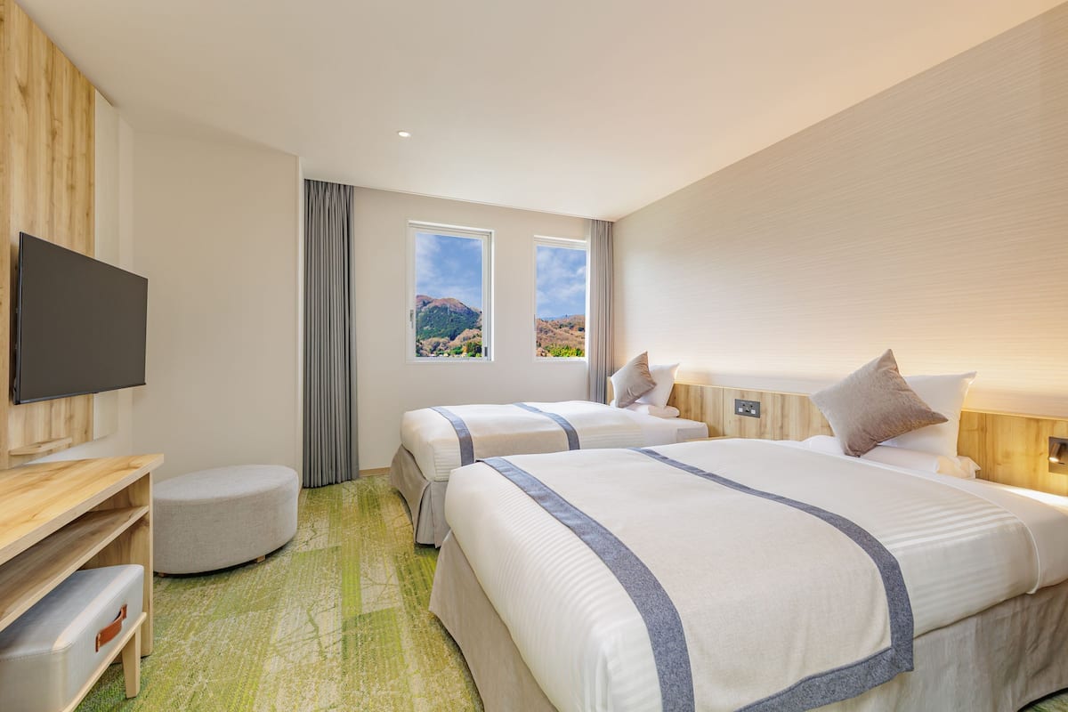 Une chambre d’hôtel avec deux lits, un téléviseur à écran plat, un bureau en bois et une vue sur un paysage montagneux, visible à travers deux fenêtres.