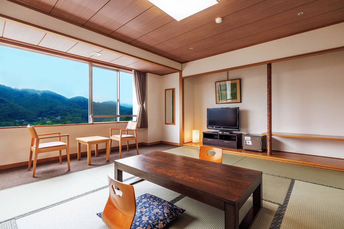 Une chambre d’hôtel avec un sol recouvert de tatamis, une table à manger en bois, des chaises, un téléviseur et une grande fenêtre donnant sur une vue sur les montagnes.