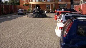 A multi-story building with a curved facade, a parking lot with several cars, and a small fountain in the center. - Great North Resort (Makuyu)
