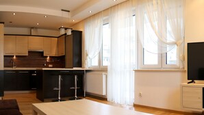 A modern living room with a flat-screen TV, a dining table, and a kitchen area with wooden cabinets and a built-in oven. - Apartament F&J 12 (Torun)